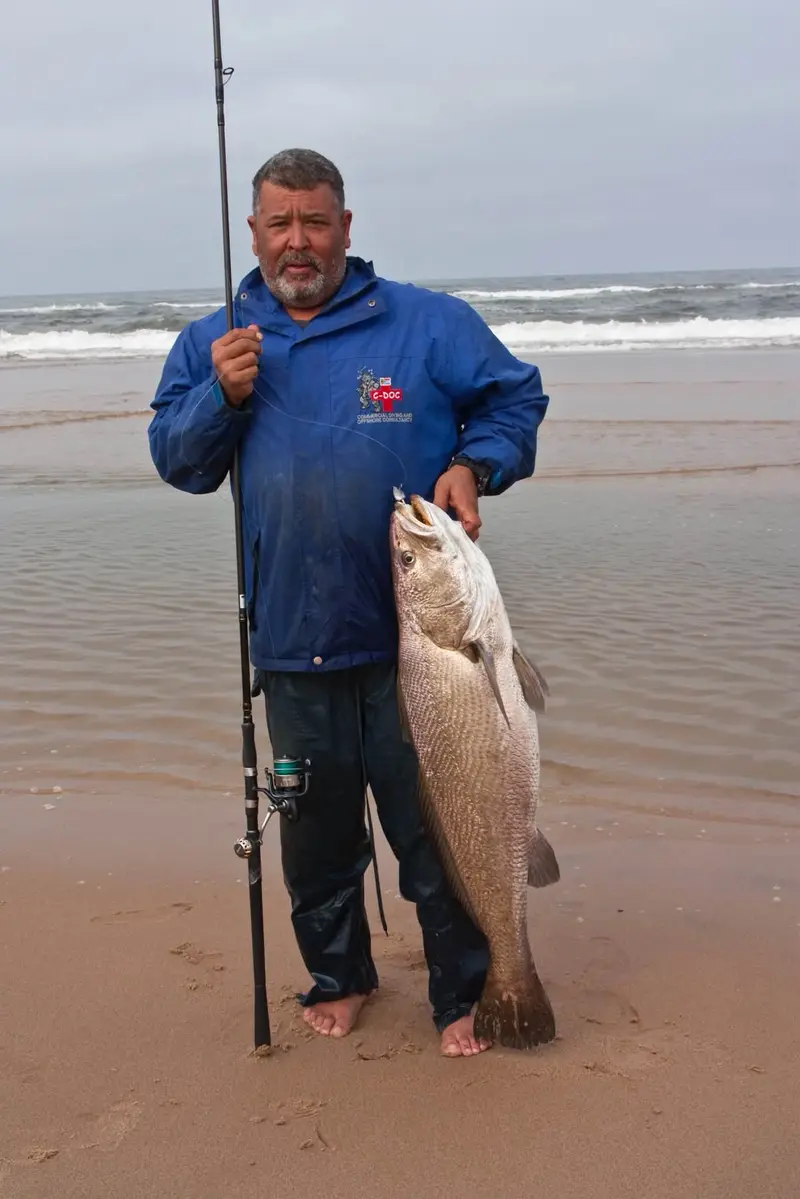 Angler in a blue jacket holding a big kob on an overcast beach