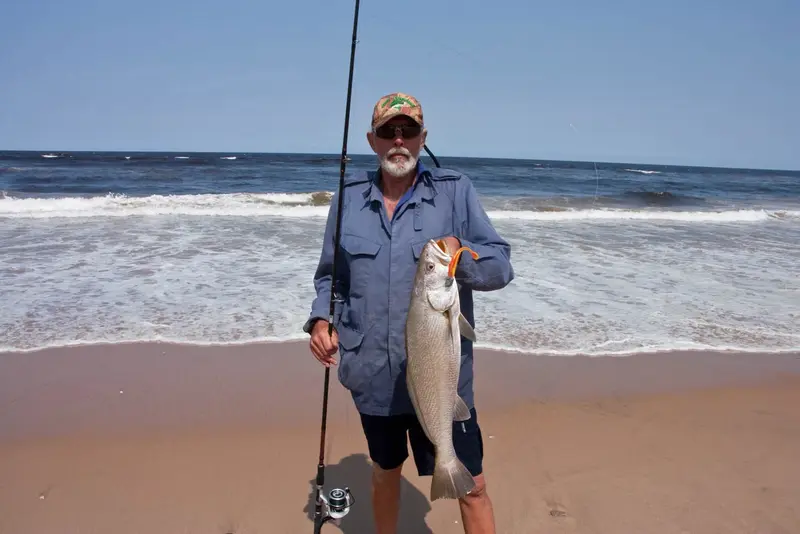 Angler in a cap holding a kob at the edge of the surf