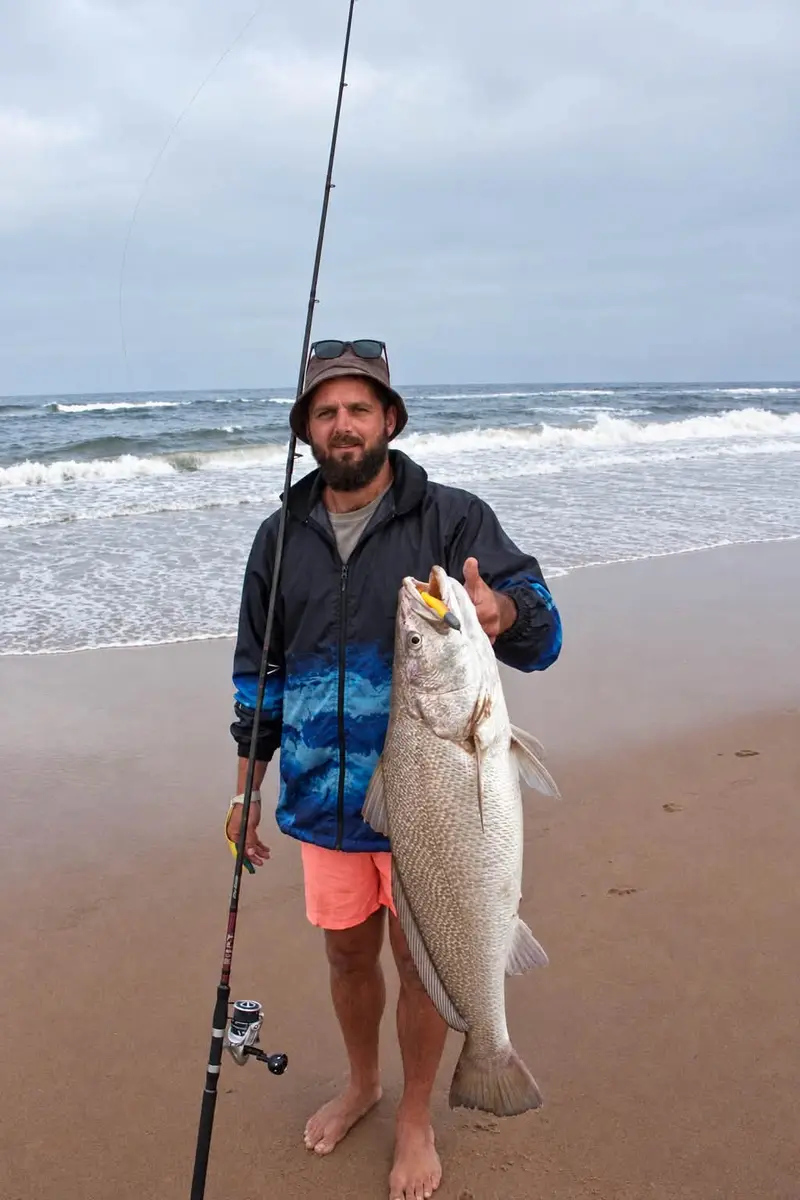 Boeta Louw in a bucket hat on an overcast beach, holding a large kob upright, rod in hand